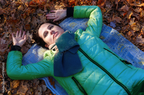 An adult motionless woman in an autumn jacket, in a scarf, lies on the ground with dry leaves, eyes open, arms bent at the elbow above her head. Up to the waist