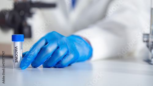 Close Up Of Female Lab Worker Wearing PPE Researching Vaccine With Microscope Holding Test Tube