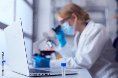 Female Lab Worker Wearing Lab Coat Working On Covid-19 Vaccine With Laptop And Microscope