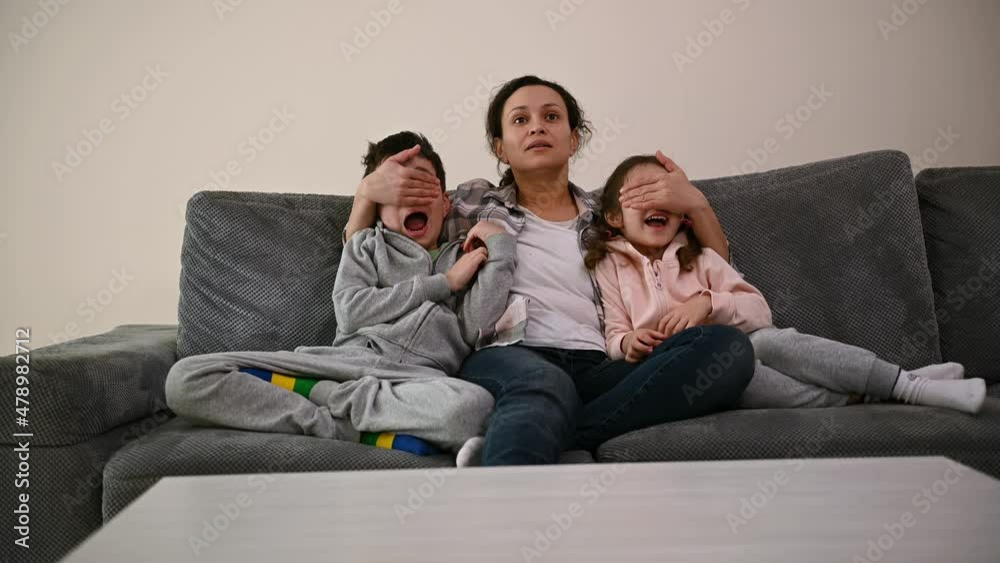Middle-aged woman, loving mother sits on the sofa and hugs her children, watches TV together when she suddenly covers their eyes with her hands Middle-aged woman, loving mother sits on the sofa and hugs her children, watches TV together when she suddenly covers their eyes with her hands
