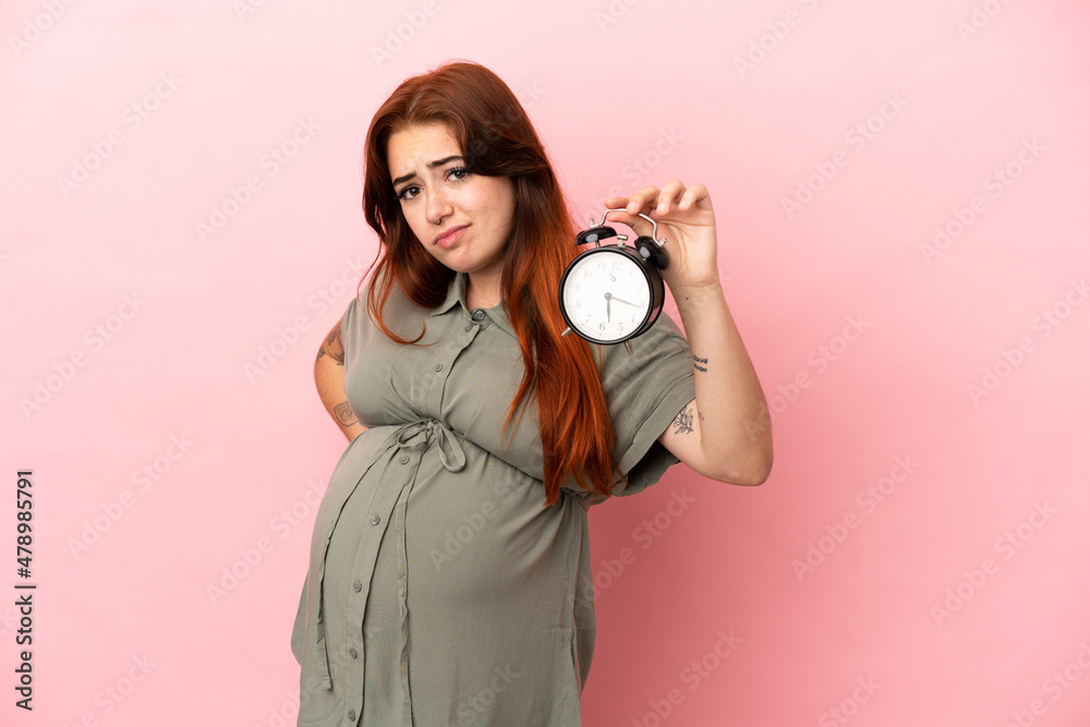 Fototapeta premium Young redhead caucasian woman isolated on pink background pregnant and holding clock while thinking