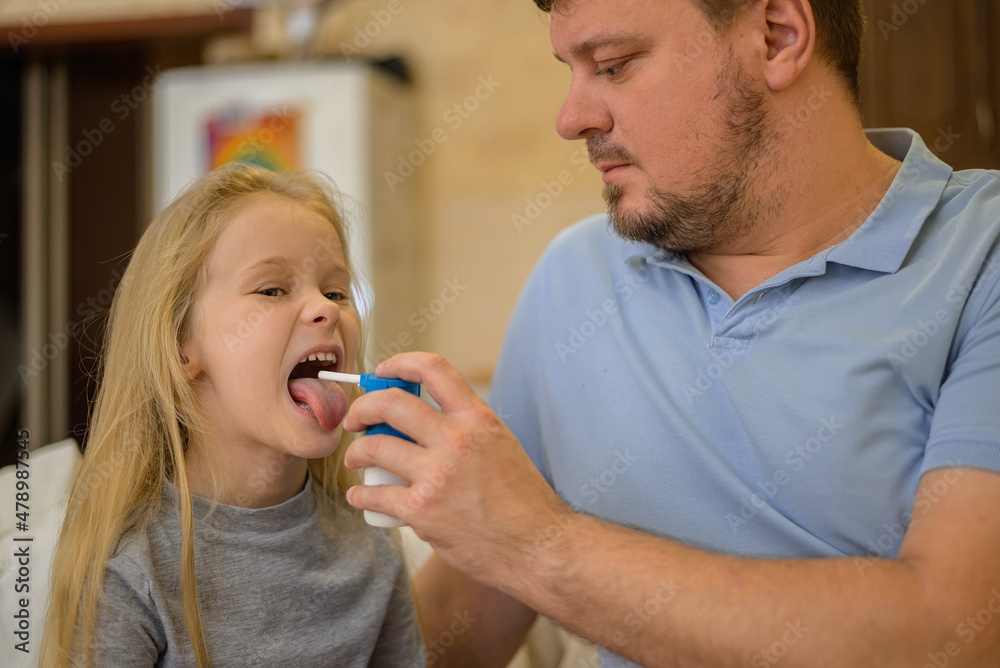 dad squirts spray medicine down the throat of his sick child on the couch at home. The father suspects that his daughter is ill. The child is not feeling well. the concept of equality in the family dad squirts spray medicine down the throat of his sick child on the couch at home. The father suspects that his daughter is ill. The child is not feeling well. the concept of equality in the family