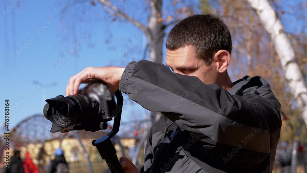 Photographer Adjusts the Camera, Camcorder on a Tripod Before Shooting Outdoors. A young male, a tourist checks the settings on professional equipment against a background of blue sky, yellow foliage. Photographer Adjusts the Camera, Camcorder on a Tripod Before Shooting Outdoors. A young male, a tourist checks the settings on professional equipment against a background of blue sky, yellow foliage.