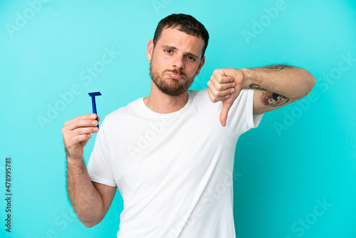 Brazilian man shaving his beard isolated on blue background showing thumb down with negative expression