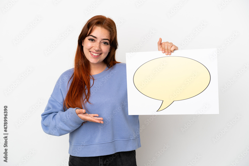 Young redhead woman isolated on white background holding a placard with speech bubble icon and pointing it Young redhead woman isolated on white background holding a placard with speech bubble icon and pointing it