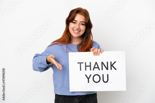 Young redhead woman isolated on white background holding a placard with text THANK YOU and  pointing it