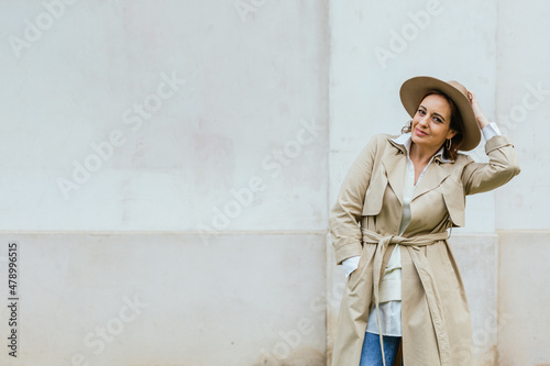 Woman in stylish trench coat and hat looking at camera while posing outdoors on the street.