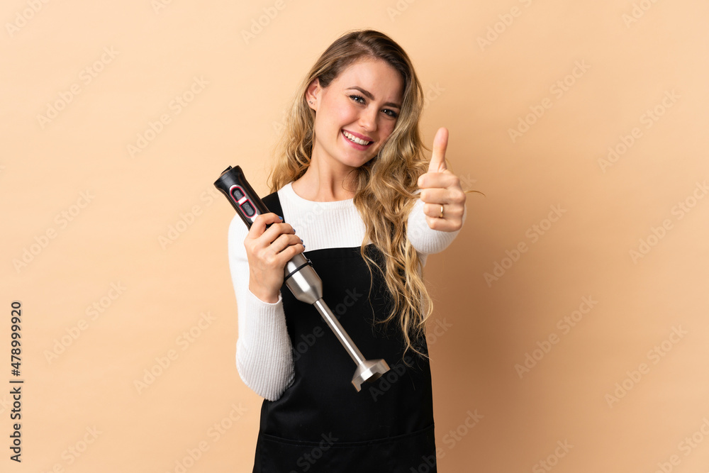 Young brazilian woman using hand blender isolated on beige background with thumbs up because something good has happened Young brazilian woman using hand blender isolated on beige background with thumbs up because something good has happened