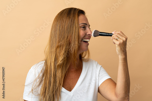 Middle age brazilian woman isolated on beige background holding makeup brush and whit happy expression