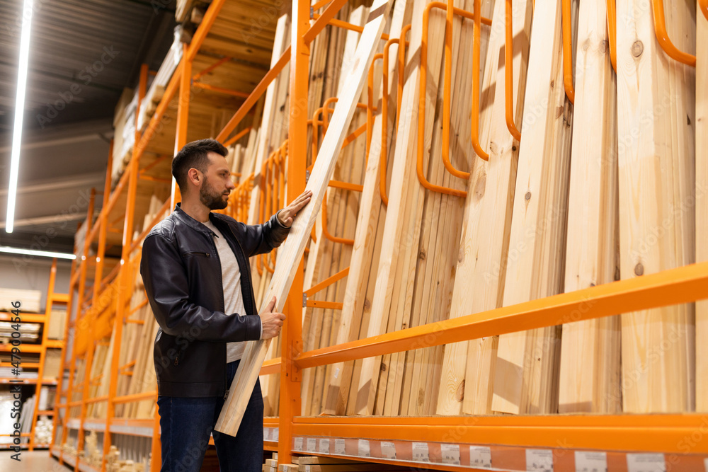 a man in a construction hypermarket in the lumber department holds a board in his hands next to a rack with wooden bars and clapboard for wall and facade decoration a man in a construction hypermarket in the lumber department holds a board in his hands next to a rack with wooden bars and clapboard for wall and facade decoration