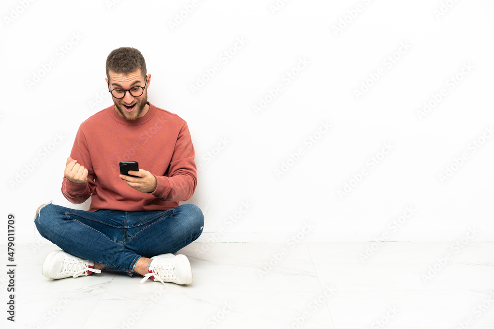 Young handsome man sitting on the floor surprised and sending a message Young handsome man sitting on the floor surprised and sending a message