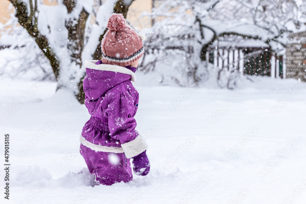 Small girl in purple overall, brown hat and purple gloves standing in deep white snow at snowy winter day. Young girl have stopped walking in snow and are looking something in distance.