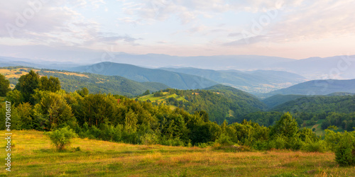 Fototapeta Naklejka Na Ścianę i Meble -  mountainous rural landscape at sunset. wonderful nature scenery with forested rolling hills and green grassy meadows in warm evening light. village in the distant valley. fluffy clouds on the sky