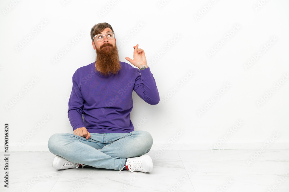 Young caucasian reddish man sitting on the floor isolated on white background with fingers crossing and wishing the best Young caucasian reddish man sitting on the floor isolated on white background with fingers crossing and wishing the best