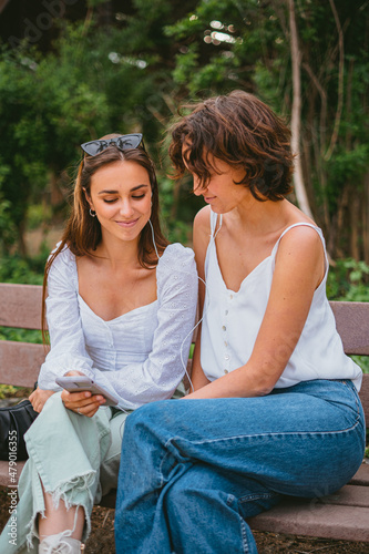Two teenage friends looking at the phone and listening to music while sitting on a bench at the park.