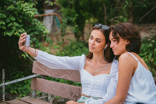 Two teenage girls taking a selfie while sitting on a bench at a park.