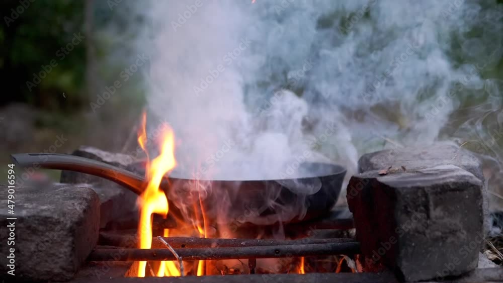 Steaming Skillet on a Blazing Bonfire in the Woods. Flaming, burnt out, frying pan in a fire, standing on a metal grate. Smoke, soot, fog over a bright flame evening. Outdoor cooking. Camping. Hiking. Steaming Skillet on a Blazing Bonfire in the Woods. Flaming, burnt out, frying pan in a fire, standing on a metal grate. Smoke, soot, fog over a bright flame evening. Outdoor cooking. Camping. Hiking.