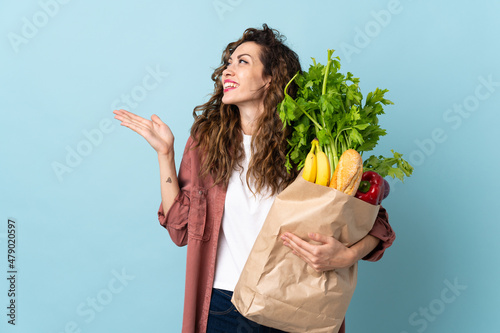 Young woman holding a grocery shopping bag isolated on blue background extending hands to the side for inviting to come
