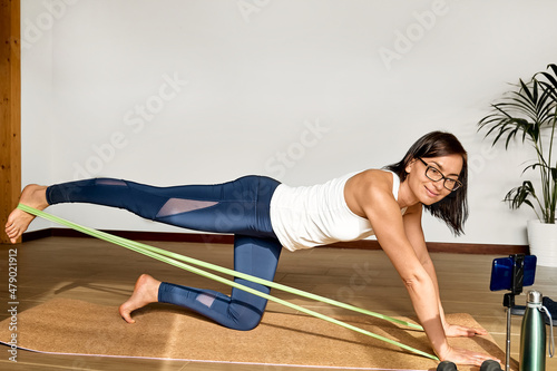 Athletic woman doing fitness training at home with resistance band following exercises online with her smartphone. Healthy lifestyle for wellness.