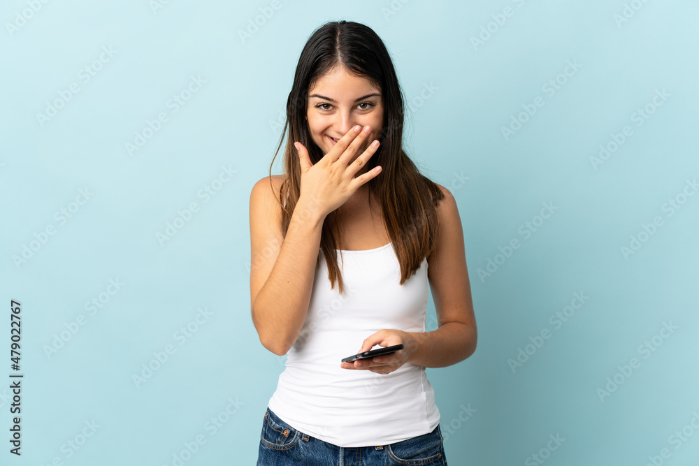Young caucasian woman using mobile phone isolated on blue background happy and smiling covering mouth with hand Young caucasian woman using mobile phone isolated on blue background happy and smiling covering mouth with hand
