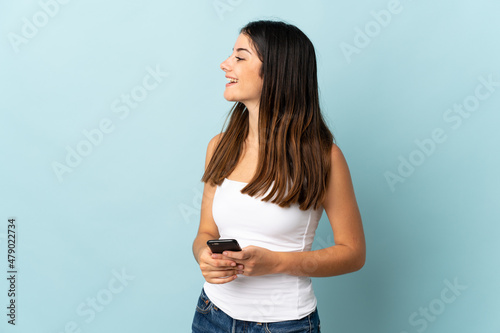 Young caucasian woman using mobile phone isolated on blue background laughing in lateral position