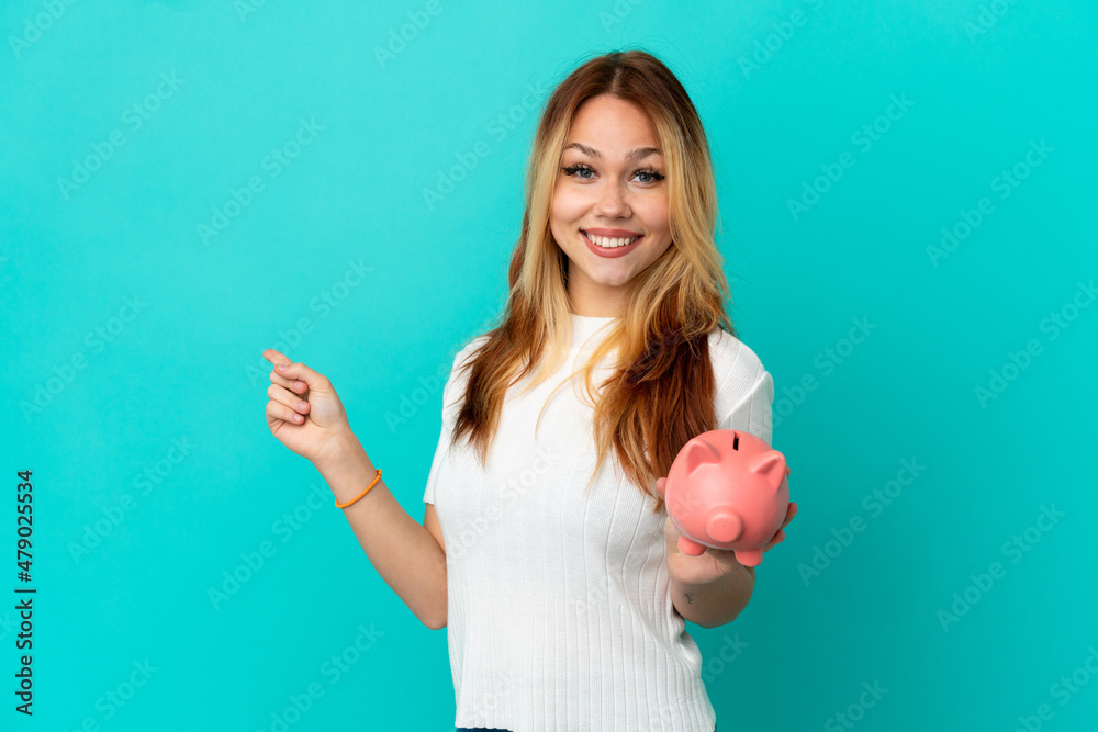 Teenager blonde girl holding a piggybank over isolated blue background pointing back Teenager blonde girl holding a piggybank over isolated blue background pointing back
