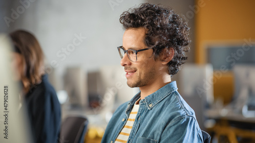 Portrait of a Handsome Smart Male Student, Studying in University, Fondly Smiling. He Wears Glasses and Has Dark Curly Hair. Works on Computer in College. Applying His Knowledge and Skills. © Gorodenkoff
