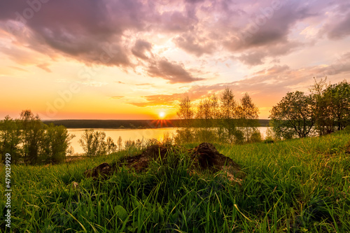 Amazing view at beautiful sunset or sunrise on a shiny lake from hill with green bushes, golden sun rays, calm water ,deep blue cloudy sky and forest on a background