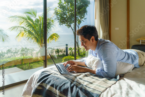 Young Asian businessman lying in bed and working with laptop in room at resort near sea during a summer vacation holiday travel.. © ake1150
