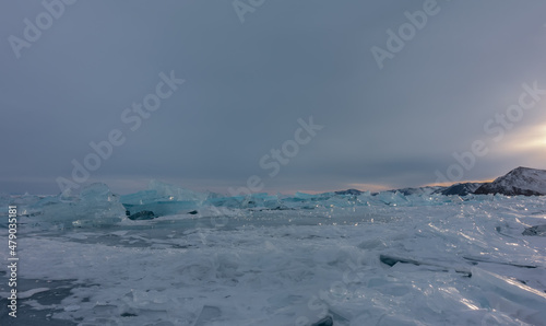 Turquoise ice hummocks are scattered in disorder on the surface of the frozen lake. The glare of the setting sun on the edges. A mountain range against the background of an evening cloudy sky. Baikal
