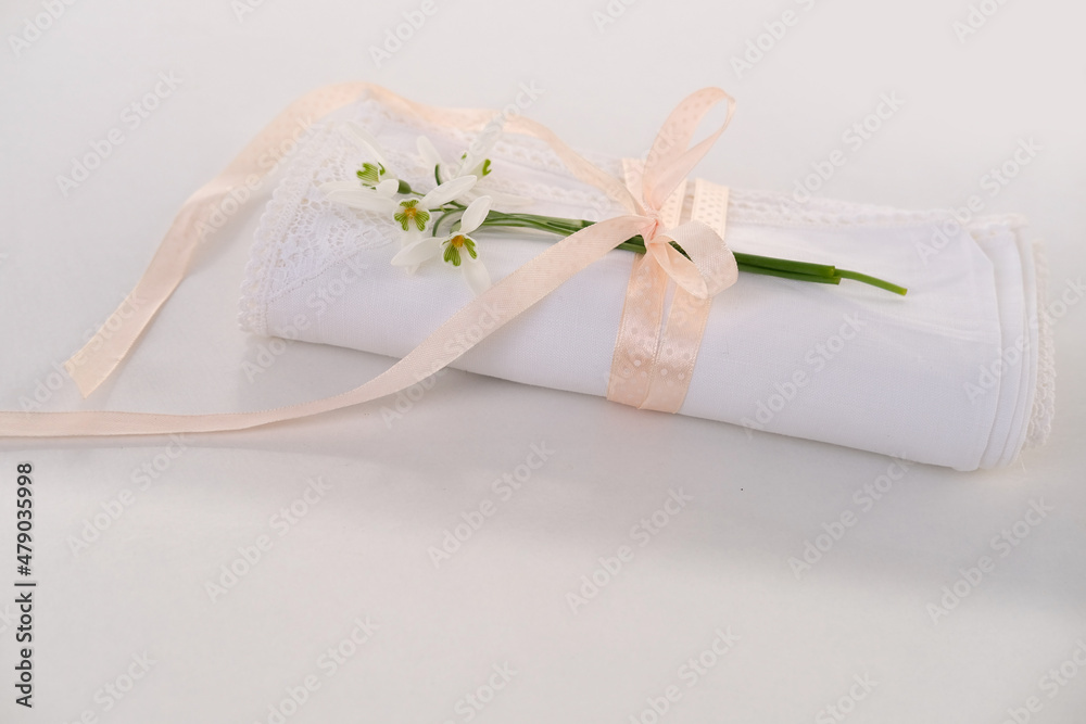 white linen napkin, finished with handmade lace, rolled in several layers, a delicate snowdrop flower is on a light table, the concept of luxury setting, a spring gift white linen napkin, finished with handmade lace, rolled in several layers, a delicate snowdrop flower is on a light table, the concept of luxury setting, a spring gift