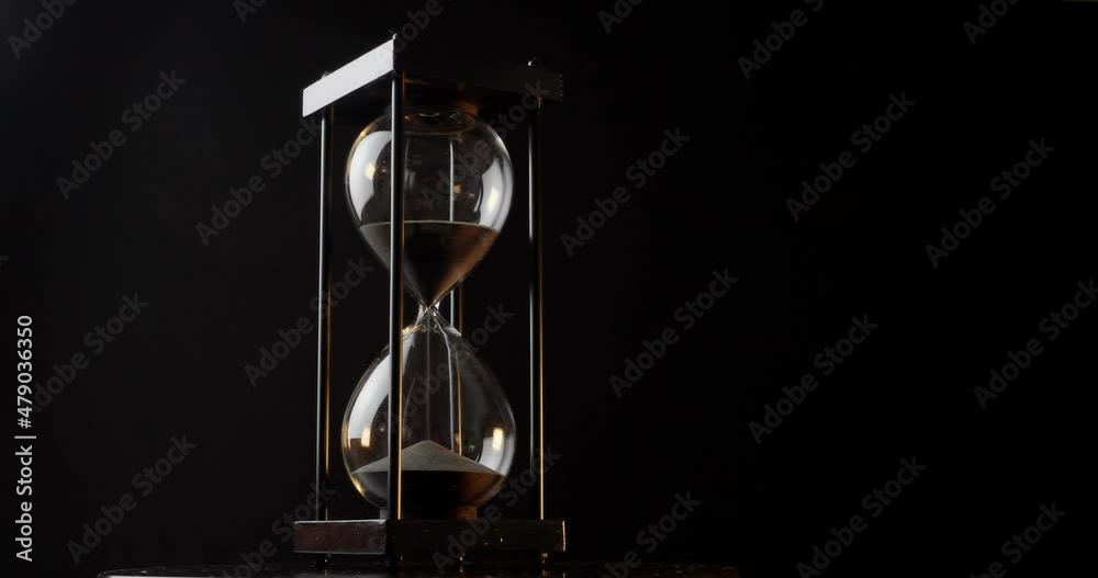 Rotation of sand timer clock with sand flowing from through hourglass on black background.
