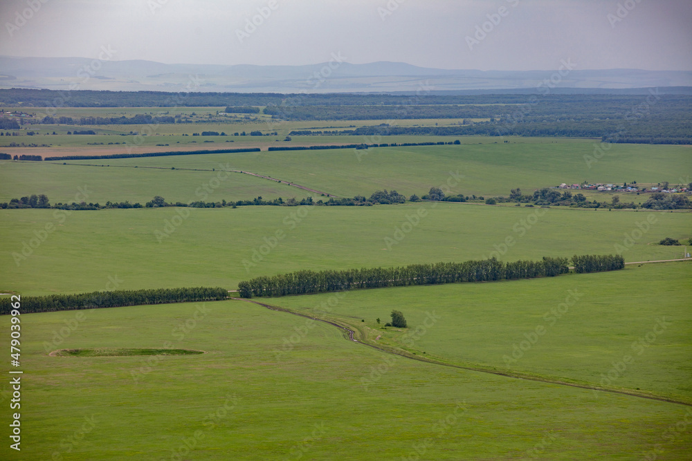 The view from the mountain to agricultural fields, separated by forest belts. Mountains on the horizon, summer cloudy landscape.
