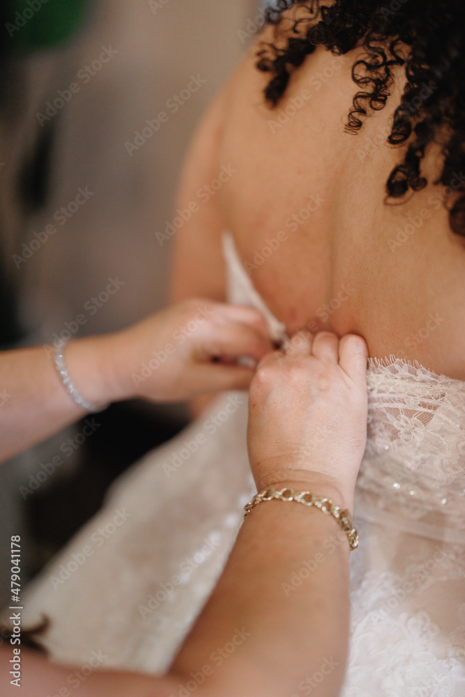 The bridesmaid helps the curly-haired bride to button up her white wedding dress Morning preparation button up the bride's dress Bridesmaid's hands fastening the wedding dress buttons Back view The bridesmaid helps the curly-haired bride to button up her white wedding dress Morning preparation button up the bride's dress Bridesmaid's hands fastening the wedding dress buttons Back view