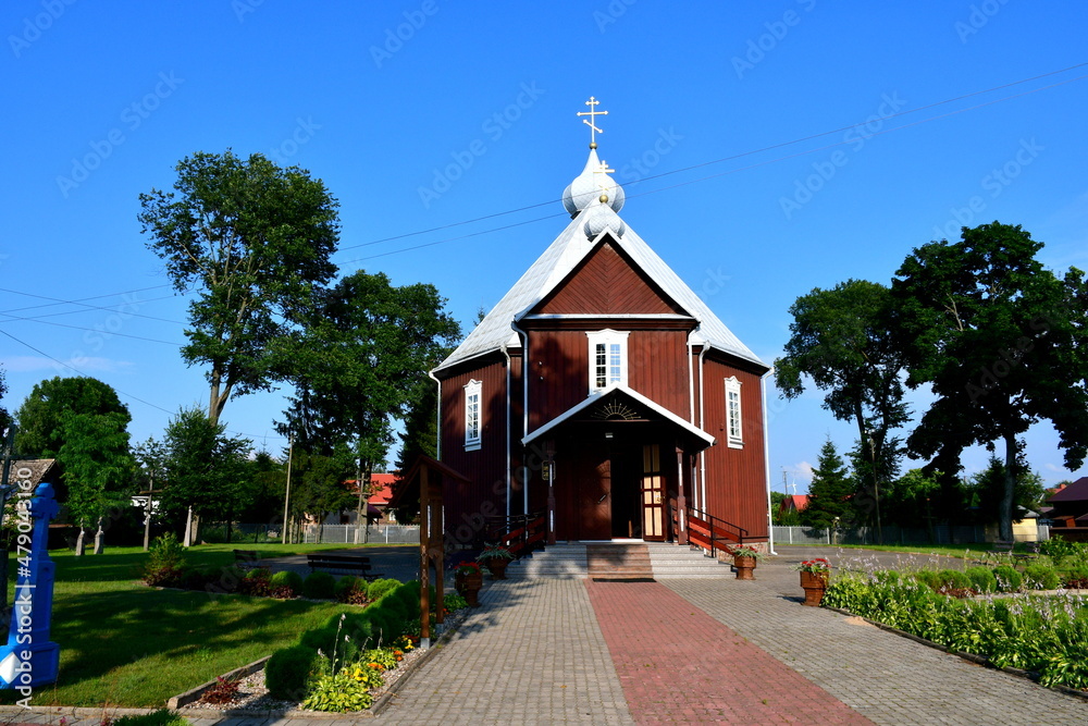 A view of an old wooden Orthodox shrine located in the middle of a well maintained public park full of shrubs, herbs, and other flora seen on a cloudless summer day on a Polish countryside