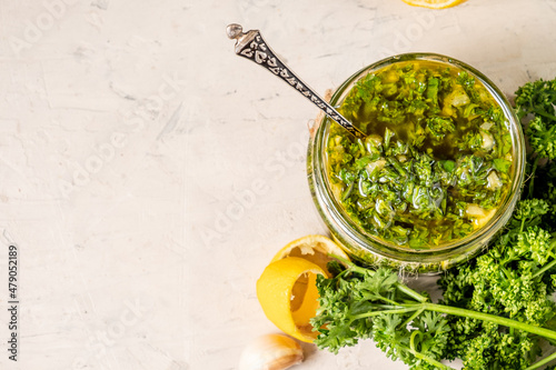 A glass jar wirh homemade fresh chimichurri sauce standing on a wooden board with herbs on a background