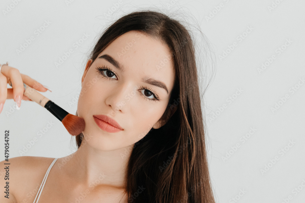 Serious young woman in the process of making makeup using cosmetic brush, posing on white studio background. Morning routine concept, getting ready for university