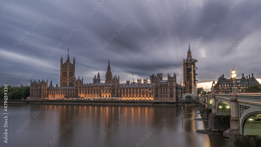 The Houses of Parliament, London, England, illuminated as night falls and reflecting on the river Thames, with clouds streaking overhead