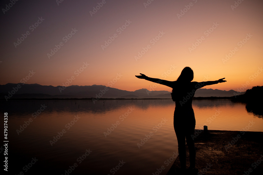 silhouette of woman opening her arms towards the horizon at sunset on pier