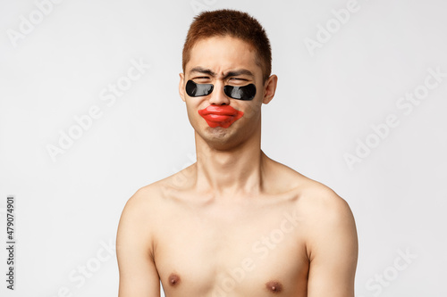 Beauty, people and lifestyle concept. Portrait of gloomy complaining funny asian boyfriend, standing naked torso and whining as girlfriend put on eye-patches and lip-patch, white background photo