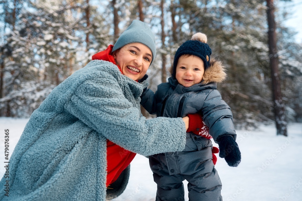 Fototapeta premium Young mother and her baby playing in the snowy forest. Young mother carrying her baby in the forest on a very snowy day.