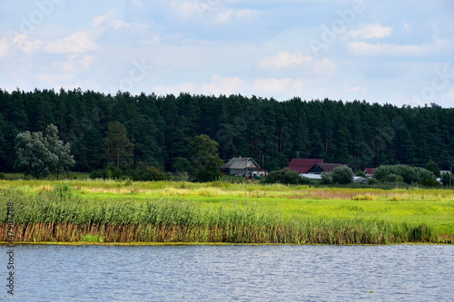 A view of a vast yet shallow lake running next to a reed covered bank or coast with some forest or moor visible in the distance next to a small rural village or cottage seen in summer in Poland