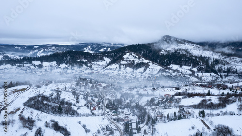 Aerial winter landscape with small village houses between snow covered forest in cold mountains