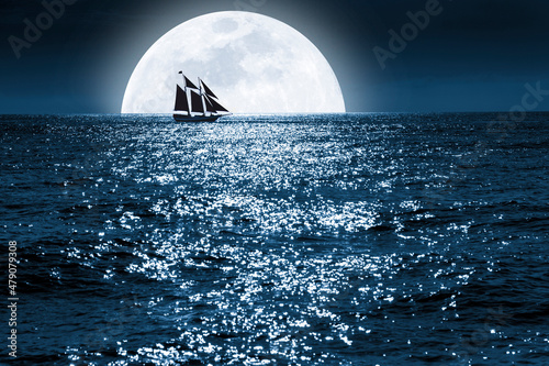 Very large full blue moon rises over this ocean scene as a sailboat quietly saiils in front of it at a distance, completely surrounded by the moonrise photo