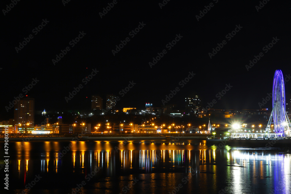 City embankment with bright lanterns and a Ferris wheel and their reflections in the water.