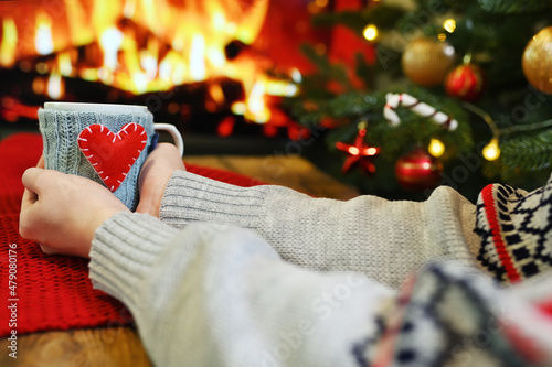 woman's hands in a warm sweater holds a cup with a hot drink in a blue knitted cover with a red heart against the background of a burning fireplace