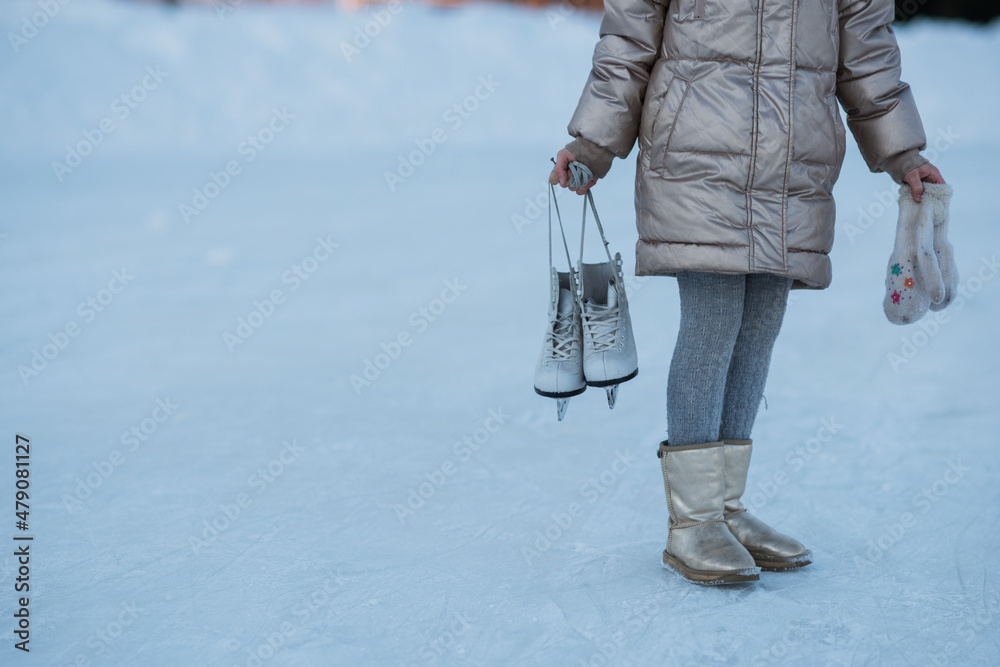A little girl stands on the ice and holds white figure skates in her hands. Active weekend getaway in winter weather. Place for your text.