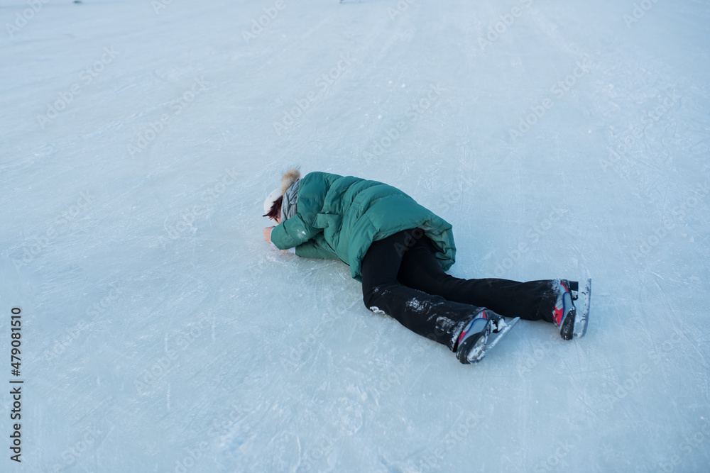 a young girl in a light-colored hat and a green jacket fell on the ice in skates, covering her face with her hands, the girl fell on the ice and was crying in pain, an injury while skating.