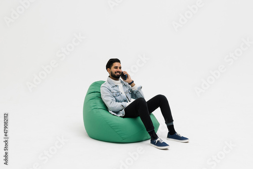 Handsome young man wearing a casual outfit, sitting on a bean bag, holding his phone talking to someone, looking away on white background
