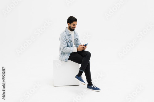 Young man sitting on chair while looking at phone celebrating on white background